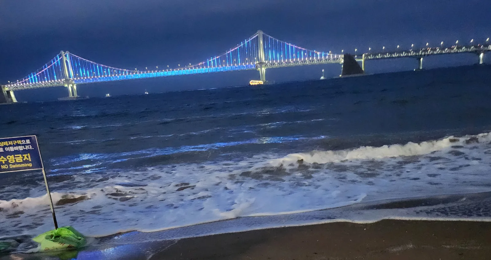 Gwangan Bridge lit in blue and pink at dusk above dark surf, with a No Swimming sign at the edge of the water