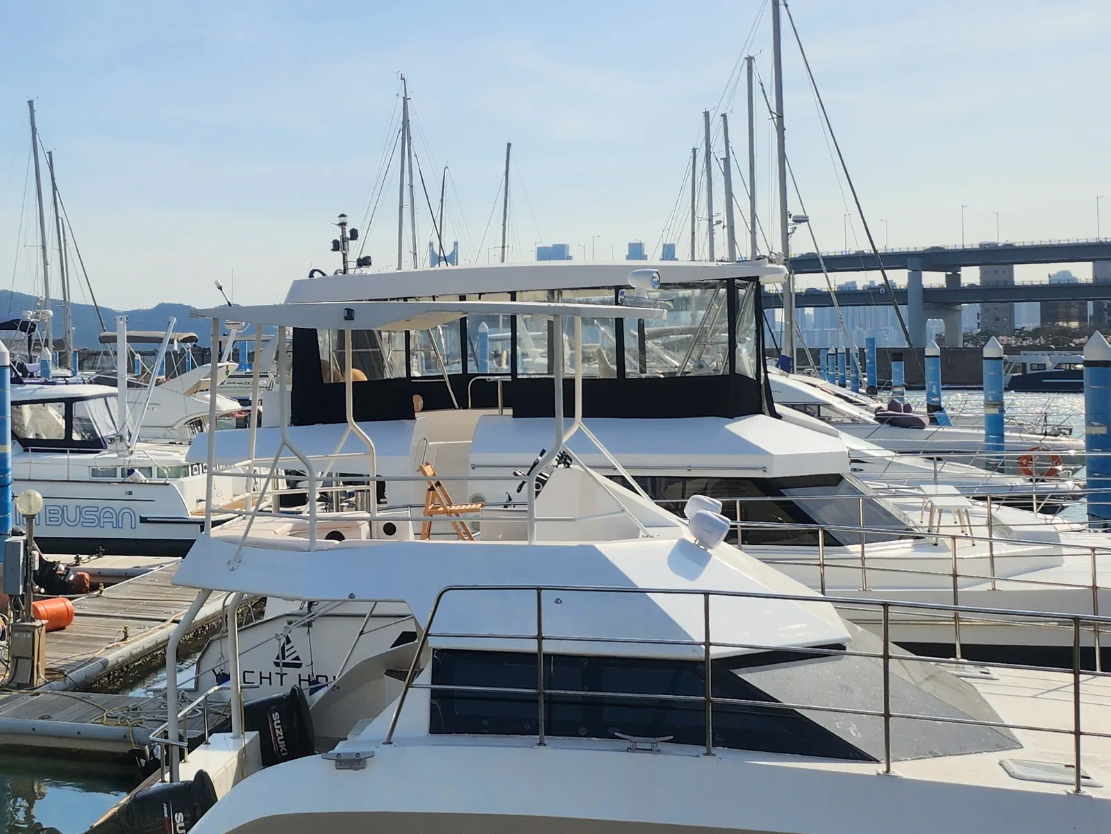 White motor yachts and sailing masts at floating pontoons with blue posts, and a double-deck road on tall piers behind them on the right